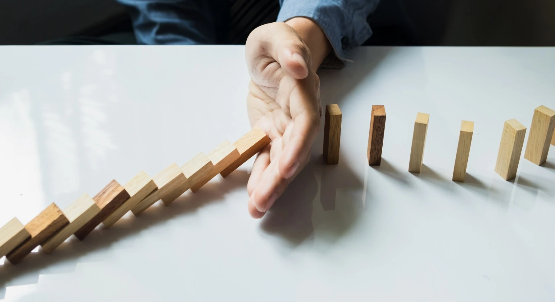 A hand interrupts a row of falling wooden dominoes.